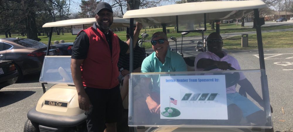Image of two men in a golf cart with a sign labeled 'Service Member Team sponsored by TIME Systems'. Lonnie Bellamy stands next to them.