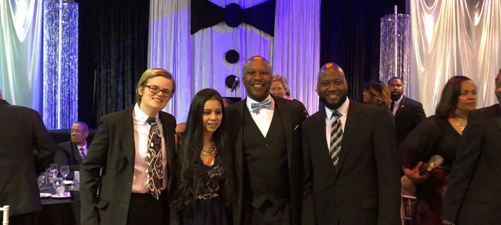 Four people in formal wear standing in front of a background resembling a tuxedo